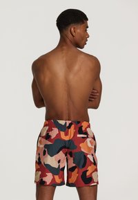 Costume de baie scurt cu talie elastică, având un model de camuflaj multicolor în nuanțe de portocalie, roz, albastru și negru, fabricat dintr-un material ușor.