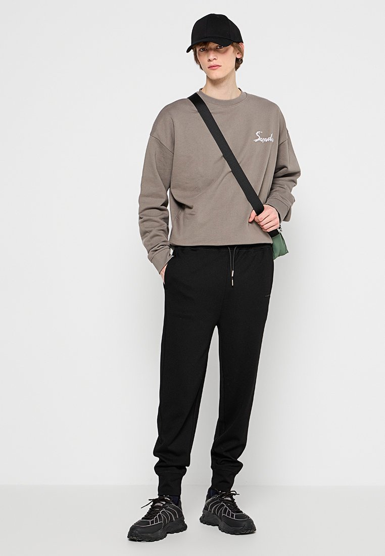 Ljusgrå kort sweatshirt med broderad text, matchad med svarta joggers. Tillbehör inkluderar en svart keps och en liten grön väska. Svarta sneakers.