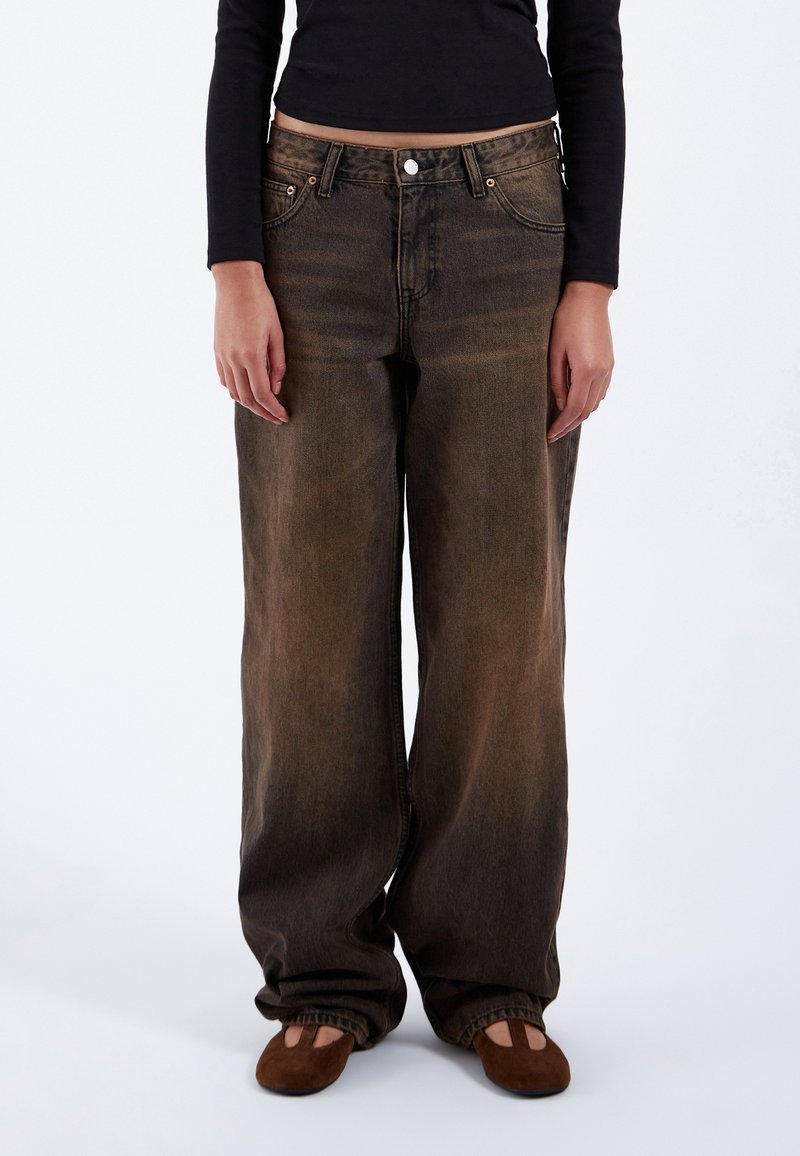 Persoon draagt losse, wijd uitlopende, vervaagde bruine jeans, bruine platte schoenen en een zwarte top met lange mouwen, staand op een witte achtergrond.
