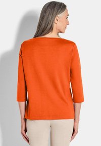 Oranger Dreiviertelarm-Top mit geradem Schnitt. Der Stoff wirkt weich und glatt und weist ein einfaches, schmuckloses Design auf.