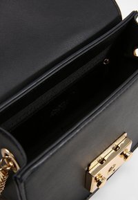 Musta nahkakäsilaukku auki, paljastaen sisäpuolen vetoketjullisen taskun ja kultasävytetyn painonapin kiinnityksen, jossa on ketjuolkaimet.