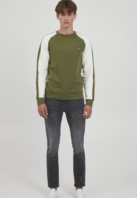 Felpa verde oliva con maniche raglan bianche, scollatura tonda e logo frontale. Abbinata a jeans neri sbiaditi e sneaker bianche.