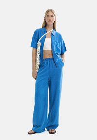 Kobieta w niebieskich szerokich spodniach, z otwartą niebieską koszulą z krótkim rękawem na białym topie, nosząca białą siatkową torbę na ramię, stoi z jedną ręką w kieszeni.