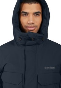 Marinblå vattentät jacka med hög krage och justerbar huva. Har två framfickor och varumärkeslogotyp på bröstet.