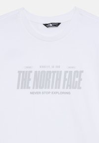 Vit bomullströja med "THE NORTH FACE" i fet grå text och "NEVER STOP EXPLORING" under, med en rundad halsringning.