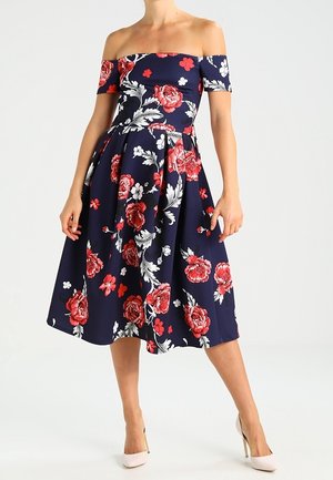 Robe mi-longue bleu marine à motifs floraux rouges, avec un décolleté épaules dénudées, une jupe évasée et un tissu lisse. Portée avec des talons clairs.