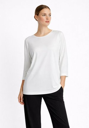 Frau trägt ein einfaches weißes Dreiviertelarmshirt und schwarze Hose, steht mit einer Hand in der Tasche und schaut zur Seite.