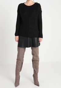 Svart stickad tröja med långa ärmar, med rund hals och knappdetaljer på ena axeln, kombinerad med en svart imitationsläderkjol och rutiga knähöga stövlar.