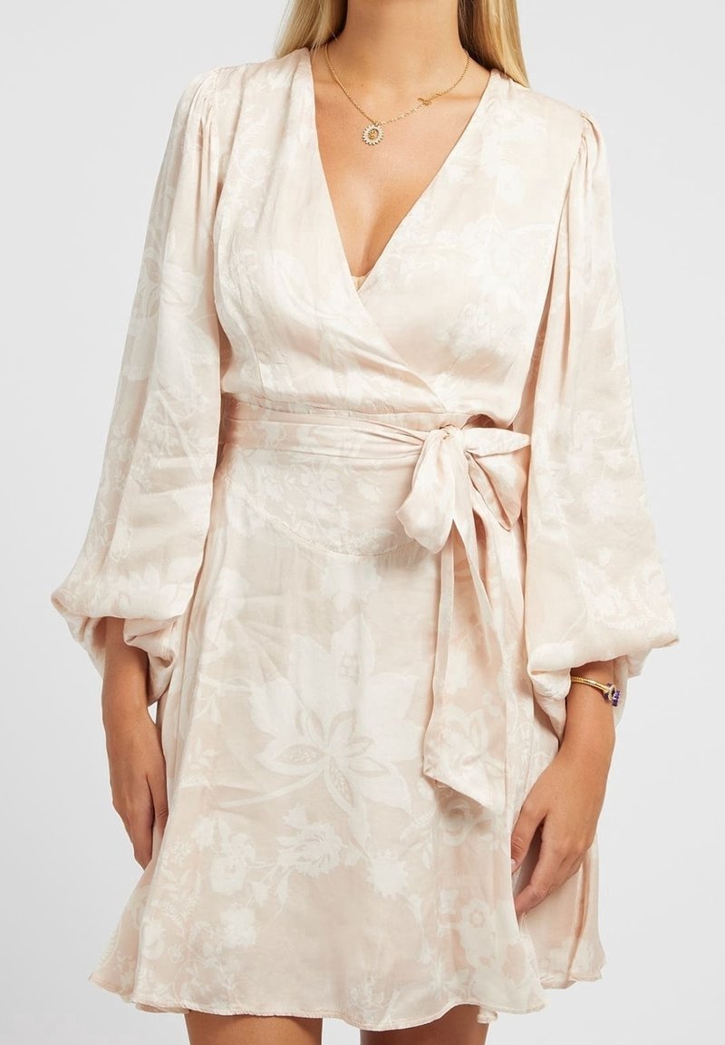 Robe portefeuille rose pâle en tissu doux, ornée de motifs floraux, avec un profond décolleté en V, des manches bouffantes et un détail de taille nouée.