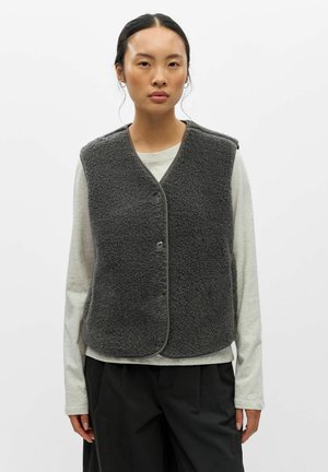 Femme aux cheveux foncés portant un gilet en polaire gris foncé sur une chemise à manches longues gris clair et un pantalon noir, faisant face à un fonds uni.