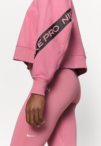 Personne portant un sweat-shirt court rose Nike Pro avec un logo sur la manche et un legging rose assorti Nike avec le logo swoosh près du genou.