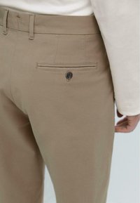 Khaki-Hosen mit glatter Textur, ausgestattet mit einer Gesäßtasche und einem Knopfdetail. Kombiniert mit einem hellen Hemd, das aus der Taille hervorschaut.