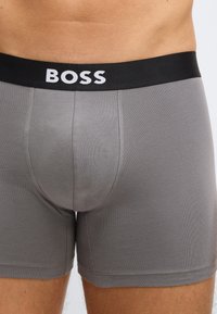 Γκρι βαμβακερά ανδρικά εσώρουχα-μπόξερ με μαύρη ζώνη μέσης που φέρει το λογότυπο "BOSS" σε λευκό. Εφαρμοστό σχέδιο με λεπτ�ή υφή και ραφές.