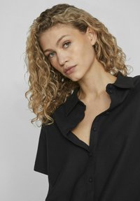 Čierna košeľa s krátkym rukávom a gombíkovým zapínaním, s golierom; vyrobená z hladkej tkaniny; má otvorený výstrih a jemné detaily gombíkov.