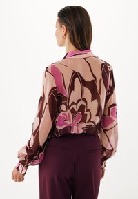 Kvinna med långt brunt hår som bär en genomskinlig blommig blus i rosa och bruna toner samt högmidjade mörklila byxor, sedd bakifrån.