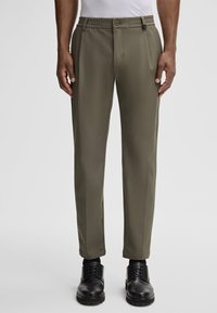 Olijfgroene broek met een gladde textuur, voorzien van een elastische tailleband, knoopsluiting en een taps toelopend ontwerp, gecombineerd met zwarte schoenen.