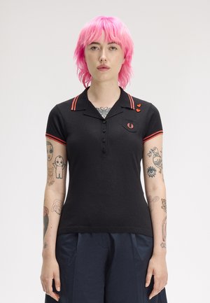 Personne aux cheveux rose vif portant une chemise noire à col avec des rayures rouges et des épingles en forme de cœur, montrant des bras tatoués, debout sur fond blanc.