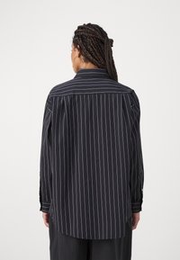Camisa negra con rayas blancas verticales, de mangas largas, cuello con solapa y dobladillo curvado. Tela suave, corte relajado y puños abotonados.
