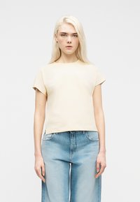 Νέος με μακριά ξανθά μαλλιά που φοράει ανοιχτό κίτρινο κοντομάνικο μπλουζάκι και ανοιχτό μπλε τζιν ψηλόμεσο π αντελόνι, μπροστά σε απλό φόντο.