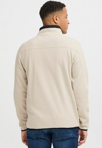 Chaqueta de forro polar beige con un cuello alto, ribete negro y acabado texturizado. Presenta costuras y un corte relajado, combinada con jeans azules.