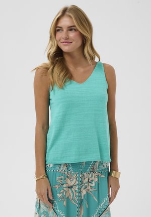 Noor naisterahvas, kellel on pikad blondid juuksed, seljas varrukateta türkiissinine särk ja lillemustriline seelik, naeratab ning vaatab veidi kõrvale.