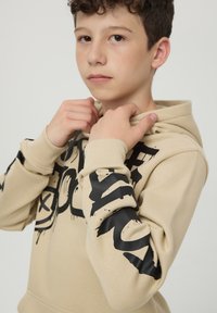 Felpa beige con stampa in stile graffiti nera sulla parte frontale e sulle maniche. Caratterizzata da cappuccio con cordino e polsini a costine. Realizzata in morbido cotone.