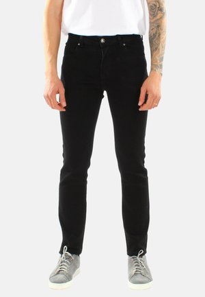 Persona che indossa pantaloni neri aderenti, camicia bianca, sneaker grigie, con avambraccio destro tatuato visibile.