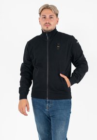 Zwarte zip-up jack gemaakt van zachte stof, met een hoge kraag, zijzakken en logo-accenten op de borst en mouw.
