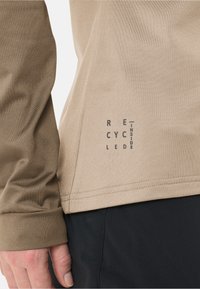 Camicia beige a maniche lunghe realizzata in materiale riciclato, con una texture liscia e una stampa con la dicitura "RECYCLED INSIDE" in lettere scure su un lato.