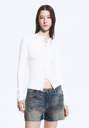 Joven mujer con cabello largo y oscuro, vestida con una camiseta blanca de manga larga abotonada y shorts vaqueros descoloridos sobre un fondo blanco liso.