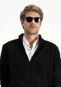 Chaqueta de forro polar negra con cremallera sobre una camisa blanca de botones, acompañada de gafas de sol oscuras. La chaqueta tiene un collar alto y un logo sutil.