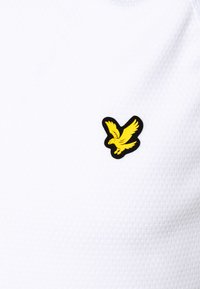 Vit polo med en texturerad tyg. Har en svart och gul örnlogotyp på vänster bröst. Enkelt design och lätt material.