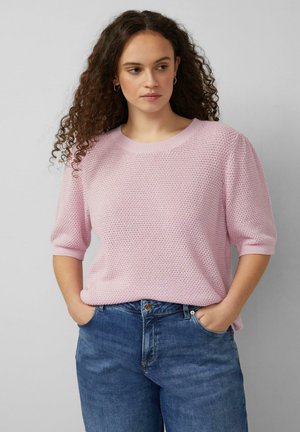 Junge Frau mit lockigem Haar, die einen strukturierten, hellrosa Kurzarmpullover und blaue Jeans trägt, die Hände in den Taschen, schaut seitlich.