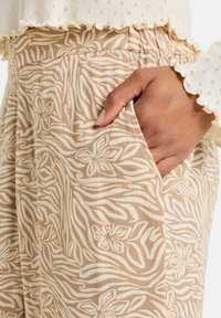 Pantaloni beige chiaro con un motivo floreale bianco e strisce zebrate, dotati di una vita elastica e tasche laterali. Tessuto texturizzato visibile.