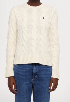 Personne portant un pull crème à torsades avec poignets et ourlet côtelés, associé à un jean bleu en denim.