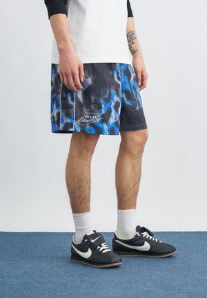 Personne portant un short Nike Air à motifs noirs, bleus et blancs, des baskets Nike noires avec un swoosh blanc, des chaussettes blanches et un haut à manches longues blanc et noir.