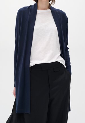 Marineblauwe lange cardigan van zachte, lichte stof, met een open voorkant, gedragen over een witte t-shirt, gecombineerd met zwarte broeken.