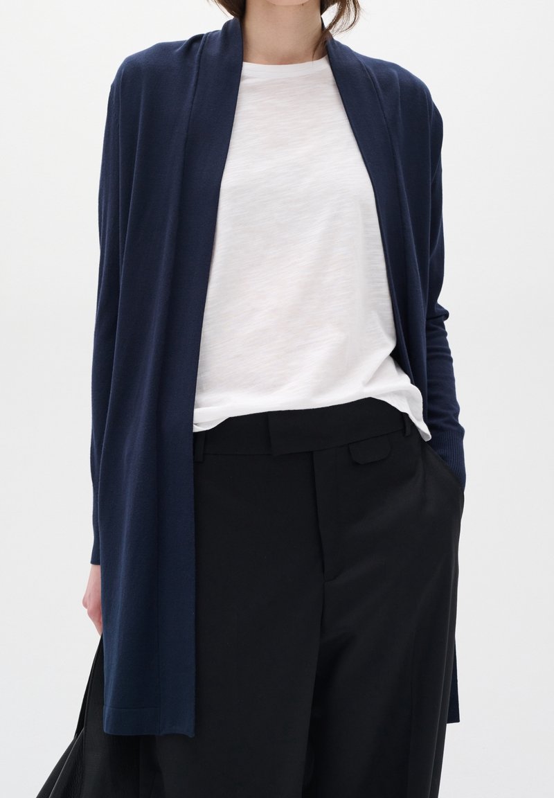 Cárdigan largo azul marino elaborado con una tela suave y ligera, diseño de frente abierto, superpuesto sobre una camiseta blanca, combinado con pantalones negros.