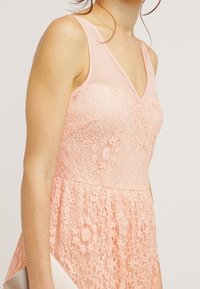Světle růžové krajkové šaty s průhlednými ramínky a přiléhavým korzetem. Krajková textura je viditelná, šaty se rozšiřují pod pasem a jsou doplněny stříbrným psaníčkem.