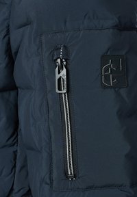 Nahaufnahme einer dunkel gepolsterten Jackentasche mit einem vertikalen silbernen Reißverschluss und einem schwarzen Gummilogo mit stilisierten Buchstaben.