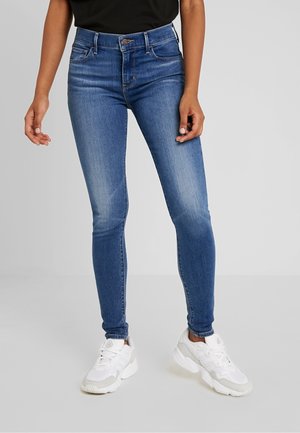 Kvinde iført blå skinny jeans med mellem talje og let falmning på lårene, kombineret med hvide chunky sneakers mod en ensfarvet baggrund.