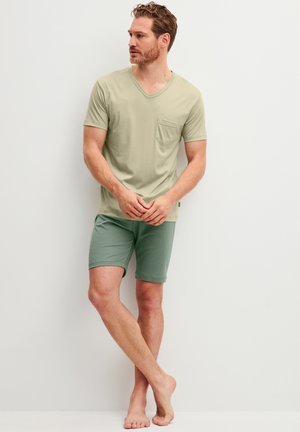Mann steht barfuß an einer weißen Wand, trägt ein beiges kurzärmeliges V-Ausschnitt-Shirt mit Tasche und grüne Shorts, schaut zur Seite.