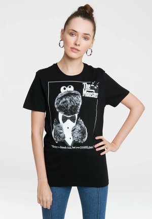 Junge Frau mit hochgestecktem Haar, die Creolenohrringe, ein schwarzes T-Shirt mit einem Smoking Cookie Monster Motiv und blaue Jeans trägt, steht vor einem weißen Hintergrund.