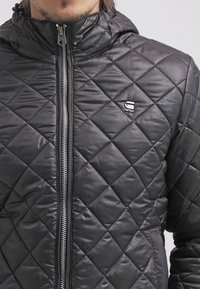 Czarna pikowana kurtka z kapturem, z zamkiem błyskawicznym z przodu i małym logo na klatce piersiowej. Materiał wydaje się błyszczący i teksturowany.