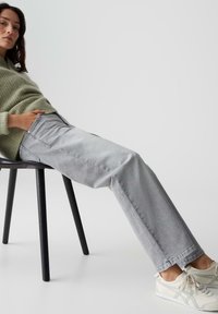 Jeans in denim grigio chiaro con taglio dritto, tasche frontali e finitura scolorita, abbinati a un morbido pullover in maglia verde chiaro e sneakers bianche.