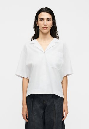 Femme aux cheveux foncés portant une chemise blanche à manches courtes avec col et un pantalon large taille haute de couleur foncée sur un fond uni.
