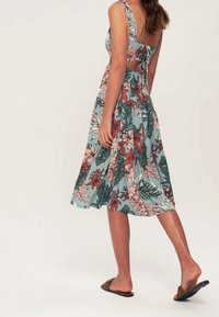 Frau in einem ärmellosen, floral bedruckten Midikleid mit offener Rückseite und Bindebändern, die barfuß in braunen Slidesandalen läuft.