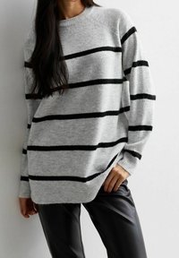 Kobieta w szarym swetrze w czarne poziome paski i czarnych skórzanych spodniach, stojąca przy gładkiej białej ścianie.