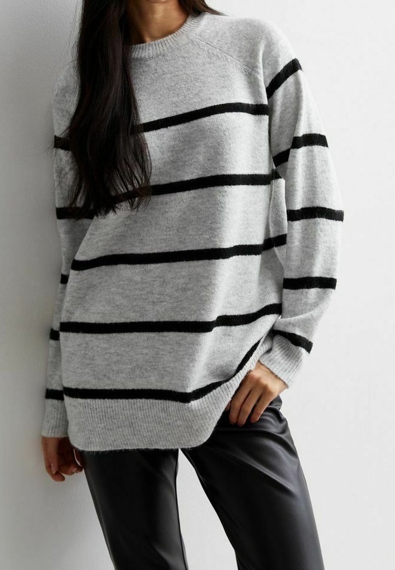 Kobieta w szarym swetrze w czarne poziome paski i czarnych skórzanych spodniach, stojąca przy gładkiej białej ścianie.