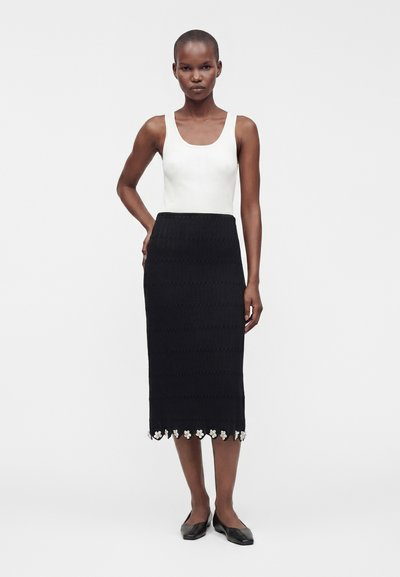 Femme aux cheveux courts portant un haut blanc sans manches et une jupe midi noire avec un bord floral, debout avec une main sur la hanche et des ballerines noires.
